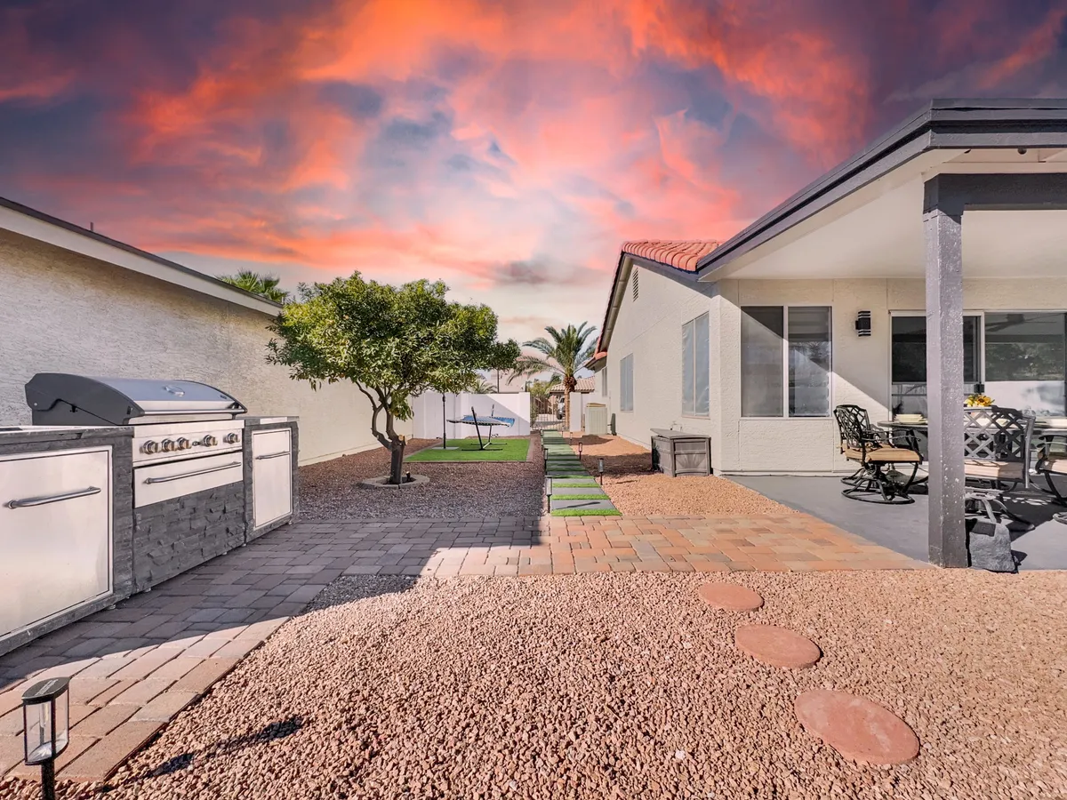 This image captures an inviting patio area with comfortable seating, a grill for barbecuing, and a landscaped path under a glowing sunset sky. The setup radiates a resort-style environment perfect for outdoor gatherings and relaxation.