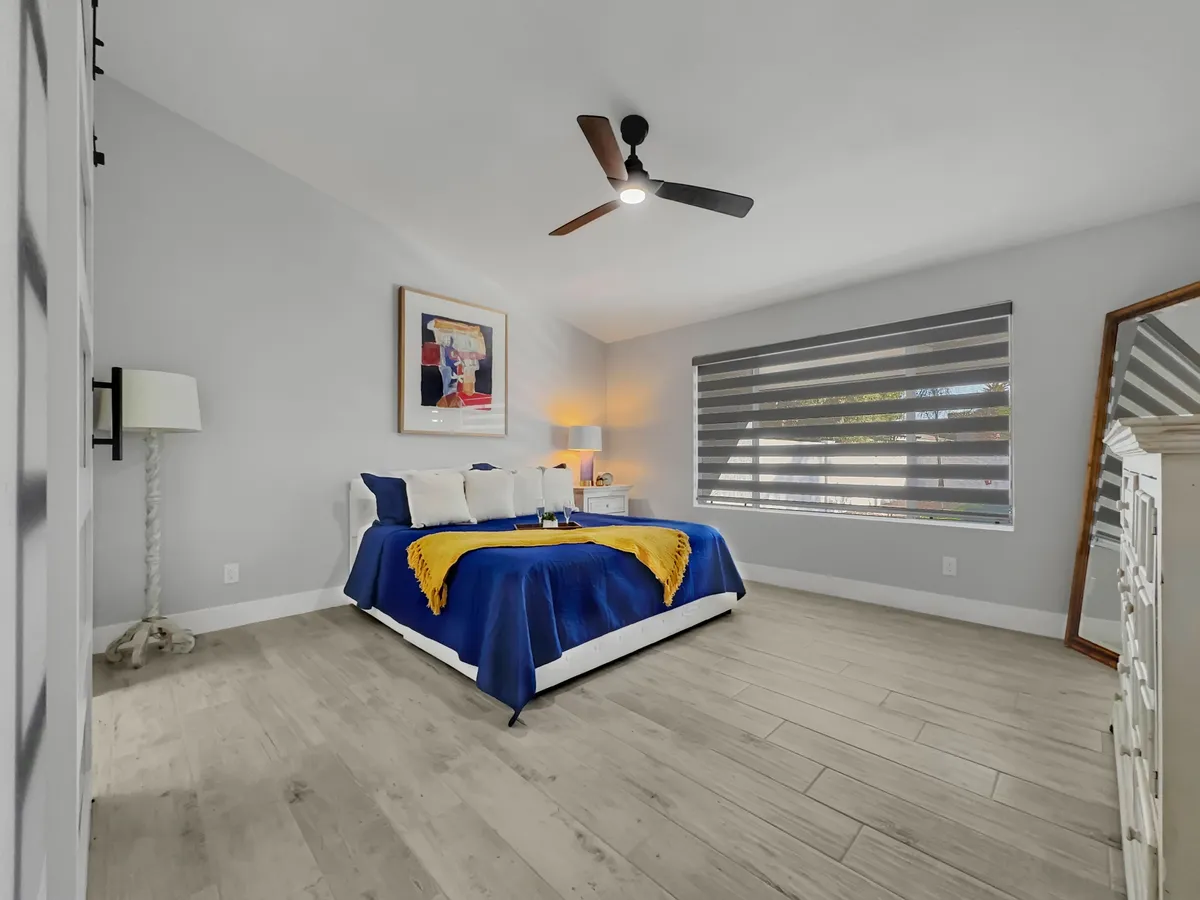 This image showcases a bedroom with blue and yellow bedding, a large window with blinds letting in natural light, a bedside lamp, and framed artwork on the wall. The mood is tranquil and cozy, ideal for relaxation and restful mornings.
