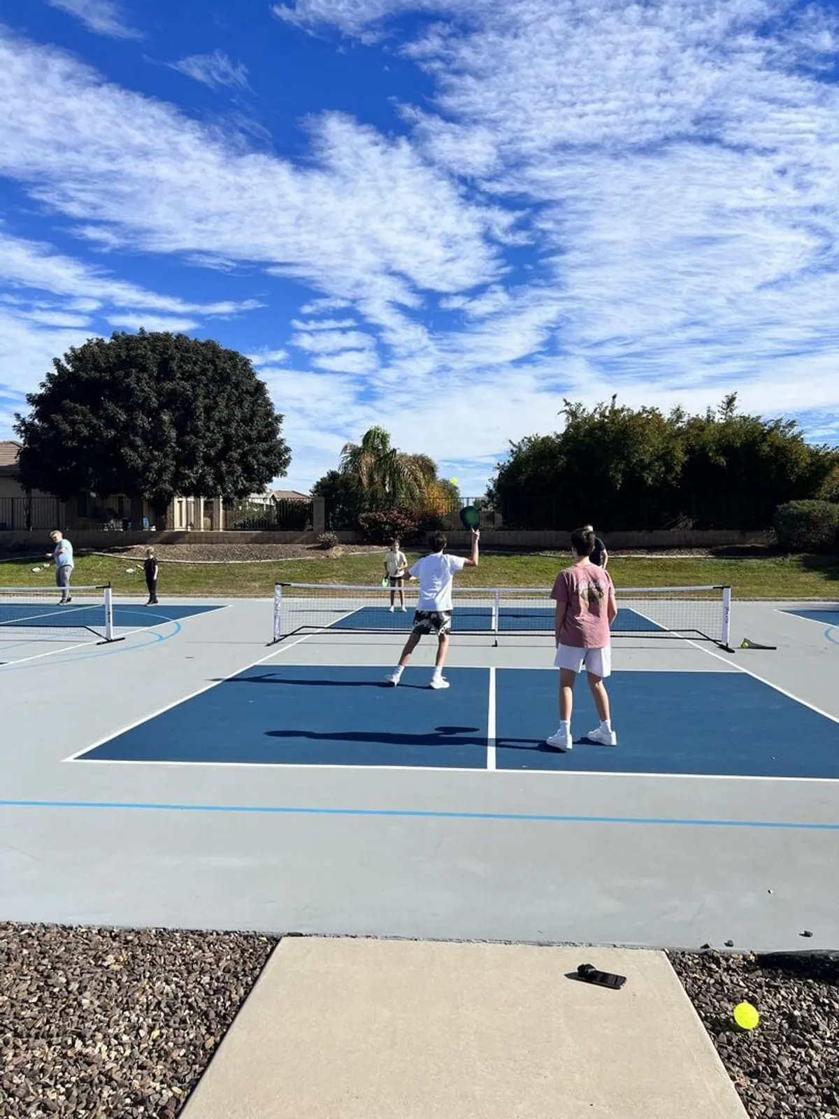 A bright outdoor tennis court with players actively engaged in a match under an open sky. The court is well-maintained and surrounded by lush landscaping, reflecting an energetic and sociable atmosphere.