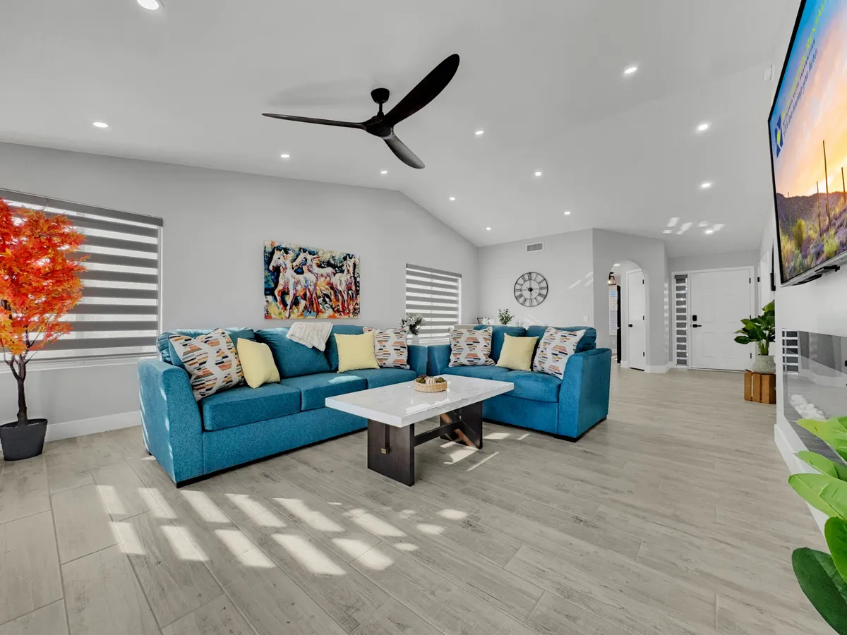 A cheerful living space highlighted by a large blue sectional sofa, a white coffee table, colorful artwork, a decorative tree with red leaves, and a modern ceiling fan. The arrangement is perfect for socializing, watching TV, or celebrating special moments with loved ones.