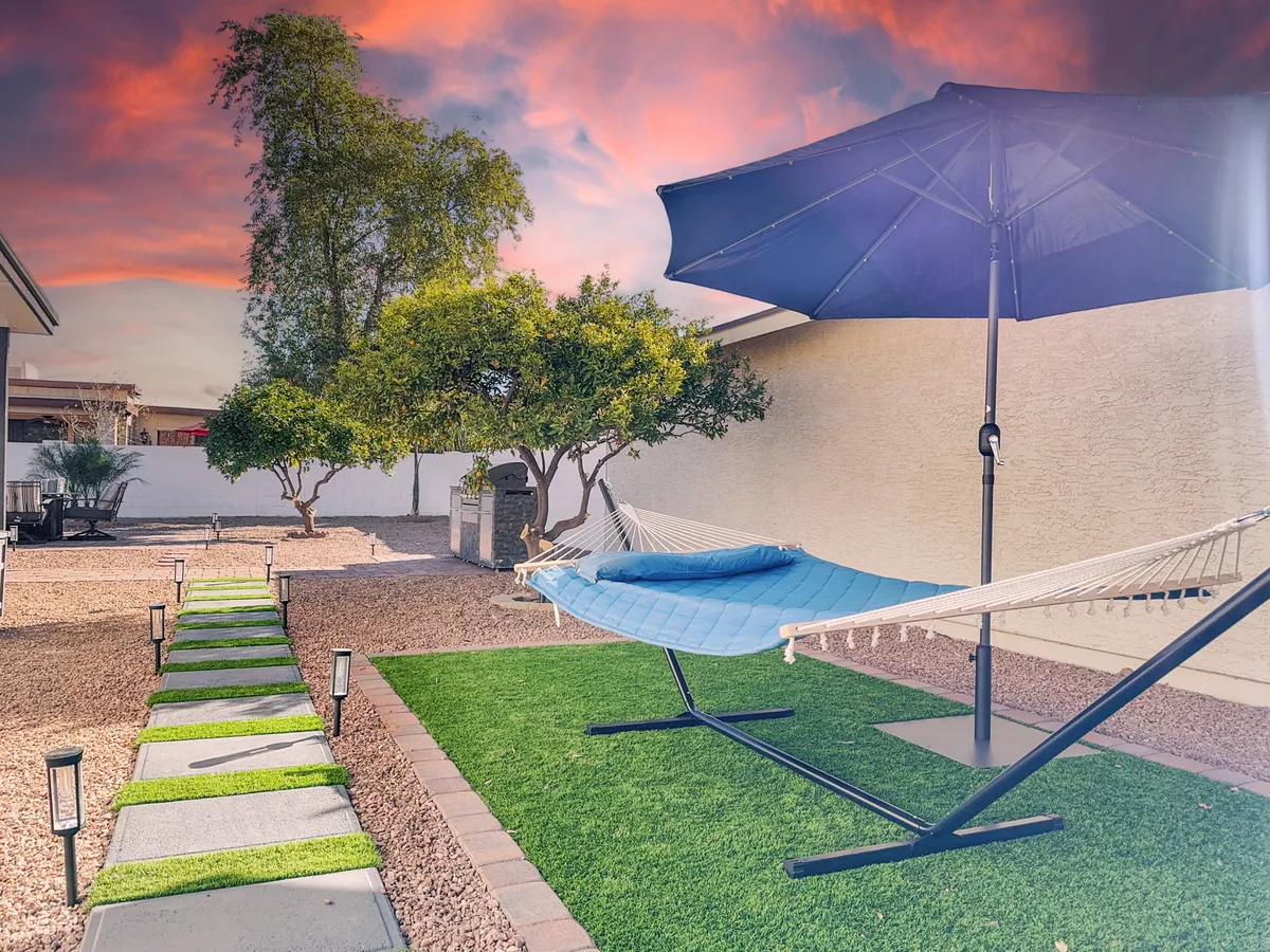 This image presents a backyard oasis complete with a hammock beneath a sun umbrella, a gently winding pathway, and lush landscaping surrounded by mature trees. The tranquil setup invites moments of relaxation, making it ideal for watching sunrises or unwinding outdoors.