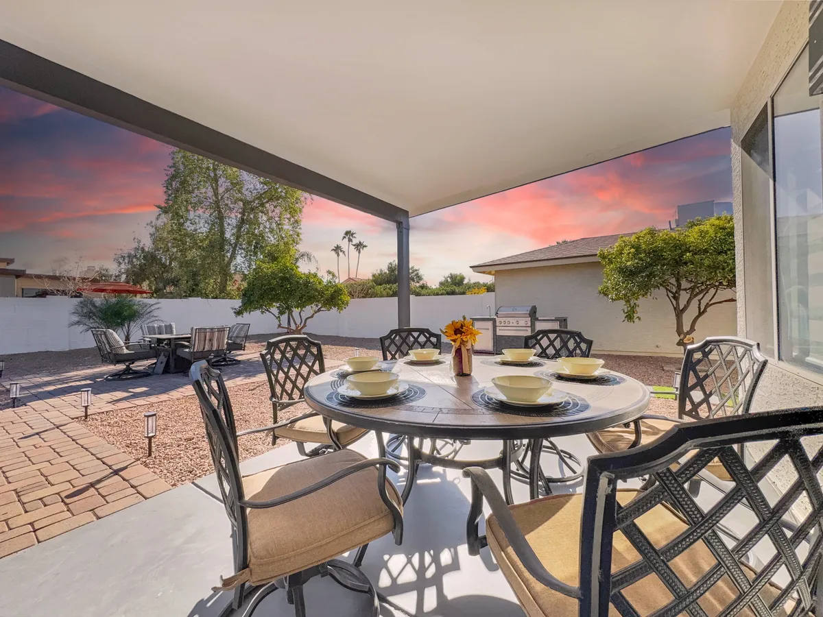 A scenic backyard or patio setting featuring a dining table adorned with fresh flowers, surrounded by lush landscaping and set against a dramatic sunset sky. The space invites gatherings, outdoor meals, and appreciation of the surrounding nature.