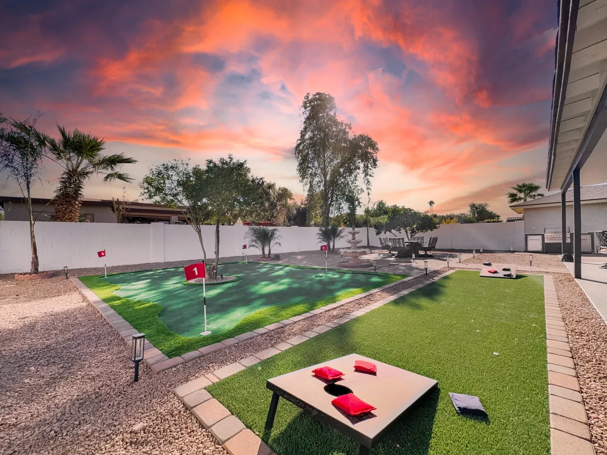The backyard scene spotlights a vibrant putting green, seating area, and visible outdoor games like cornhole, all set beneath a colorful sunset sky. This lively yet relaxed environment invites playful competition and evening relaxation.