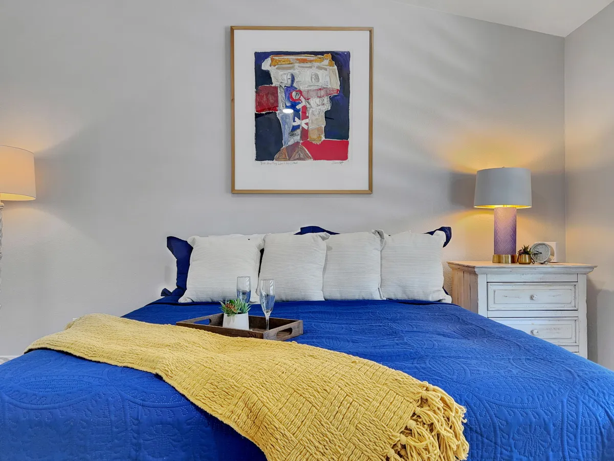 This image presents a bedroom with a blue bedspread, yellow throw blanket, artwork above the bed, and a nightstand with lamp. The space feels tranquil and comfortable, ideal for restful mornings or cozy evenings.