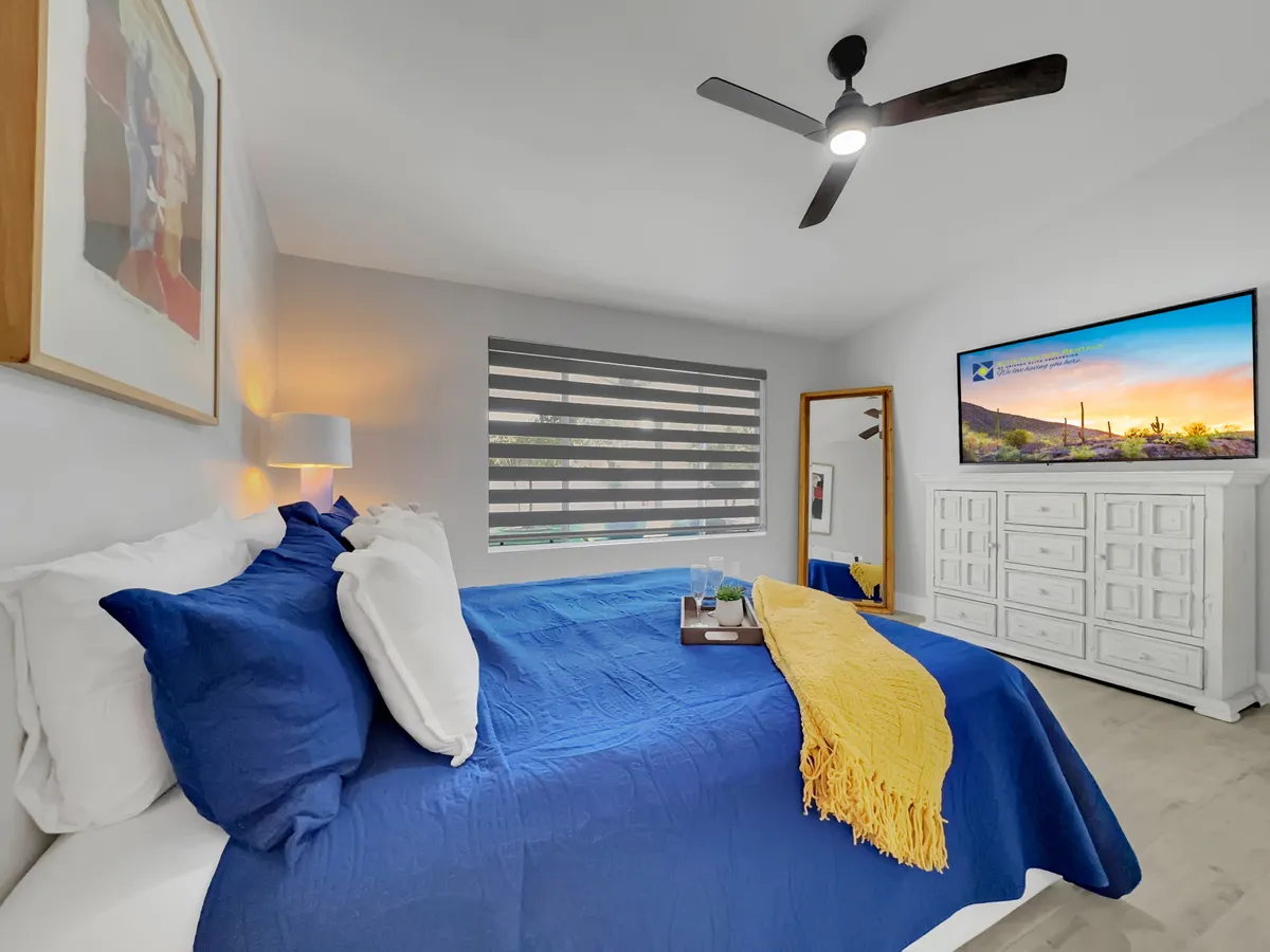 This photo features a bedroom with a blue bedspread, white dresser, framed artwork, and a TV mounted on the wall. The mood is comfortable and peaceful, suggesting a restful retreat.