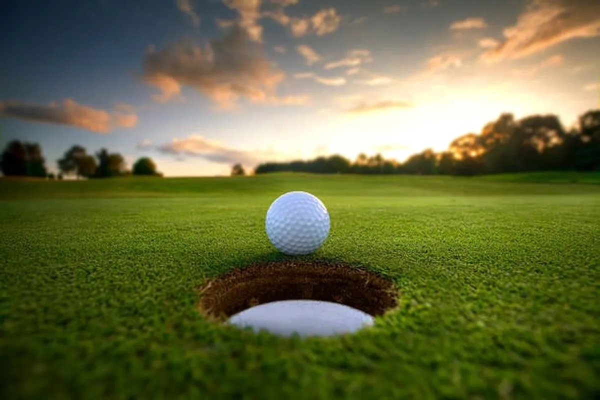A close-up of a golf ball near the hole on a lush green golf course, with ambient natural light and serene surroundings. The image evokes the experience of golfing and socializing outdoors.
